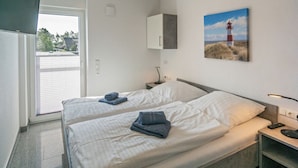 A hotel room with two beds, a small bedside table with a lamp, a painting of a lighthouse, and a sliding glass door leading to a balcony. - Holiday Home With 1 Bedroom (Esens)