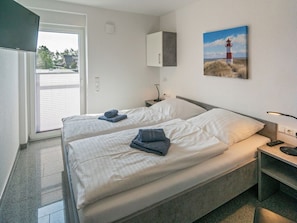 A hotel room with two beds, a small bedside table with a lamp, a painting of a lighthouse, and a sliding glass door leading to a balcony. - Holiday Home With 1 Bedroom (Esens)