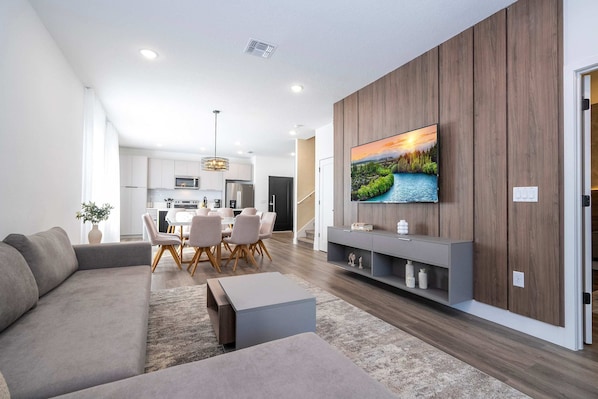 Un soggiorno moderno con una TV a schermo piatto montata su una parete in legno, un divano grigio, una zona pranzo con tavolo e sedie, e una cucina sullo sfondo.
