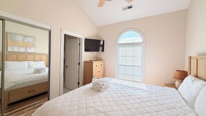 Ein Schlafzimmer mit einem Bett, einem Fernseher, einem Fenster mit Jalousien und einer Tür zu einem anderen Raum.
