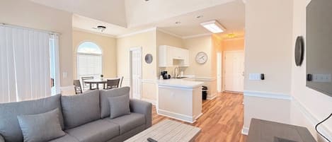 A living room with a sofa, coffee table, and remote controls. There is a dining area with chairs and a table. The kitchen is visible with cabinets and a countertop.