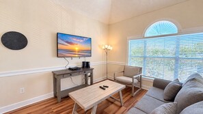 Ein Wohnzimmer mit Flachbildfernseher, Couch, Couchtisch und einem Fenster mit Jalousien.