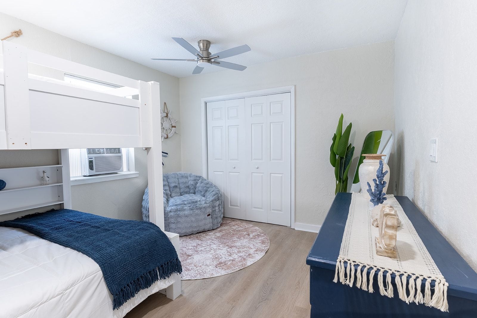 A bedroom with a bunk bed, a ceiling fan, a round rug, a desk with a vase, and a door.