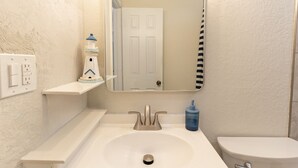 Ein Badezimmer mit einem weißen Waschbecken, einer Toilette, einem Spiegel und einem kleinen Regal mit einem Leuchtturm-Dekor.