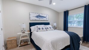 Ein Schlafzimmer mit einem Bett, Nachttischen, einem Deckenventilator, einem Fenster mit Vorhängen und einem an der Wand befestigten Bild von einem Fisch.