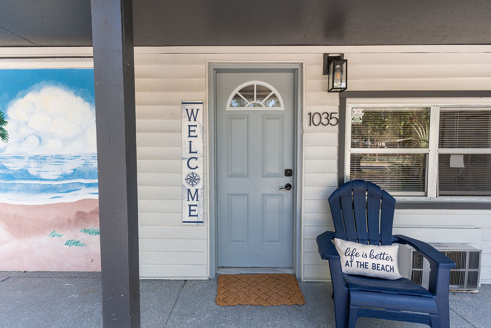 Uma casa com um letreiro 'WELCOME', o número '1035' na porta e um mural com tema praiano na parede.