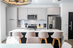 Een moderne keuken met witte kasten, een roestvrijstalen koelkast, een magnetron en een koffieapparaat. Er staan vier barkrukken met houten poten en een witte tafel ervoor.
