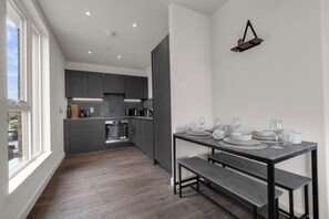 A modern kitchen with dark cabinets, a dining table set with plates and glasses, and a hanging light fixture. - Luxury Flat - Sleeps 6 - Private Balcony - 10 Min to Wembley Stadium - WiFI (Harrow)