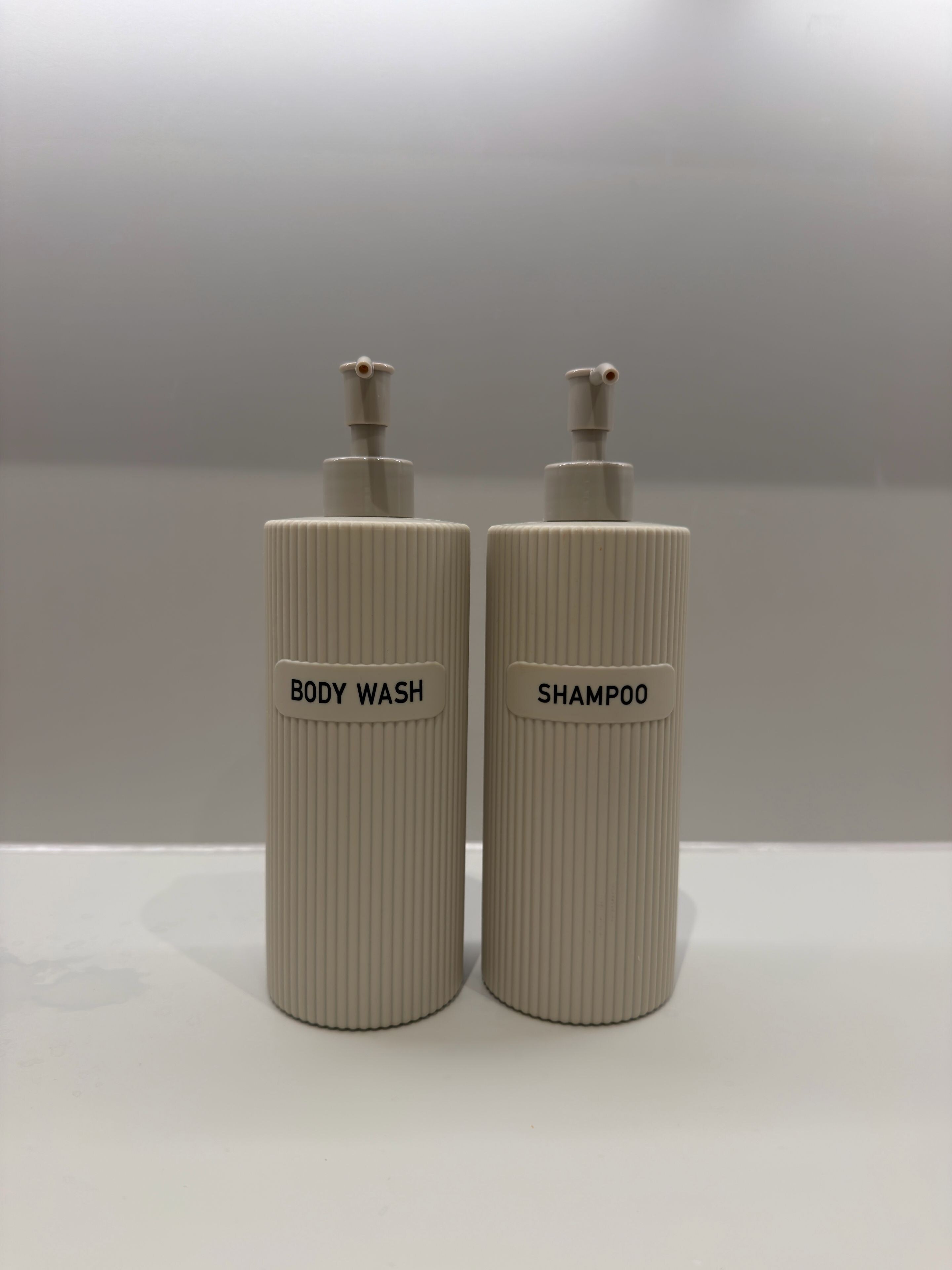 Two cylindrical bottles with pump dispensers, labeled 'BODY WASH' and 'SHAMPOO,' placed on a flat surface.