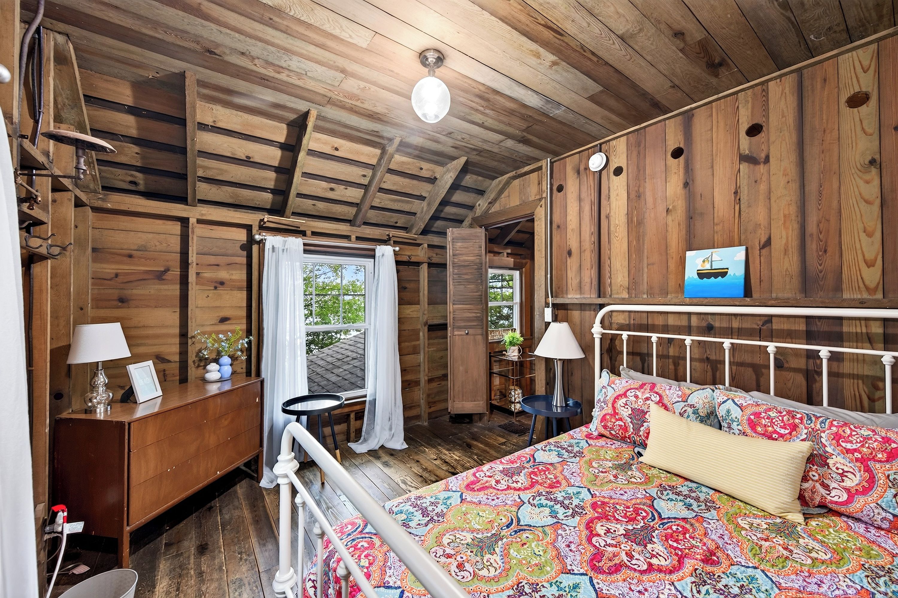 A wooden cabin room with a bed, a desk, and a window with curtains.