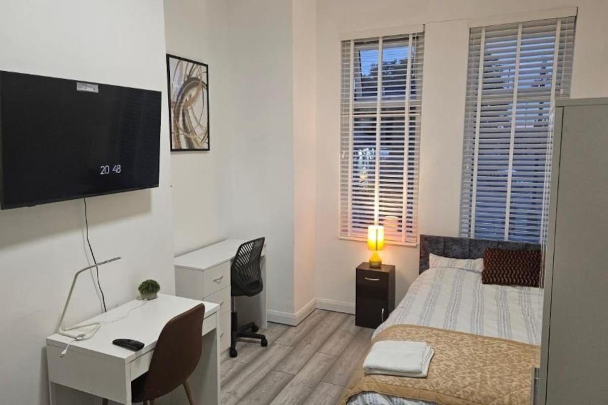Une chambre à coucher comprenant un lit, un bureau, une chaise, un téléviseur et une fenêtre avec des stores.