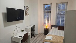 Quarto com cama, escrivaninha, cadeira, televisão e janela com persianas.