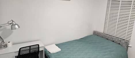 A bedroom with a bed, a desk with a chair, a lamp, and a window with blinds.