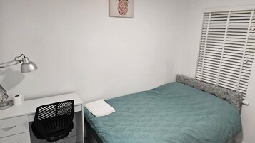 A bedroom with a bed, a desk with a chair, a lamp, and a window with blinds.