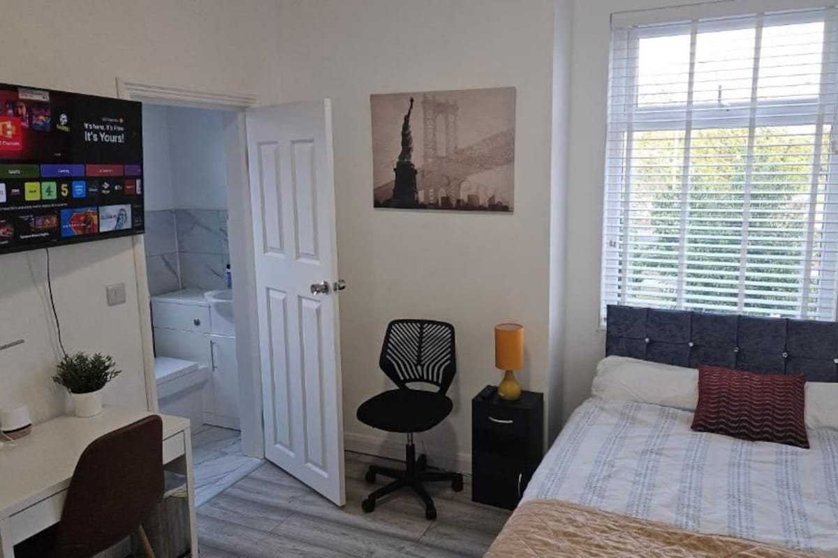 A bedroom with a bed, a desk with a computer, a chair, a window with blinds, and a door leading to another room.