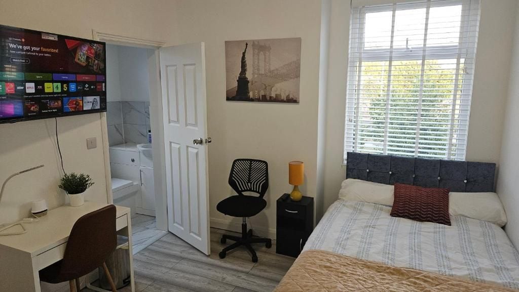 A bedroom with a bed, a desk, a chair, a TV, a door, and a window.
