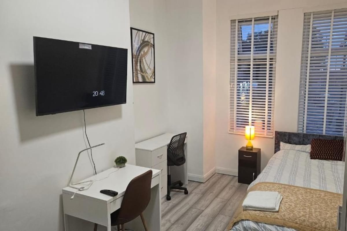A bedroom with a bed, a desk with a chair, a television, and a window with blinds.