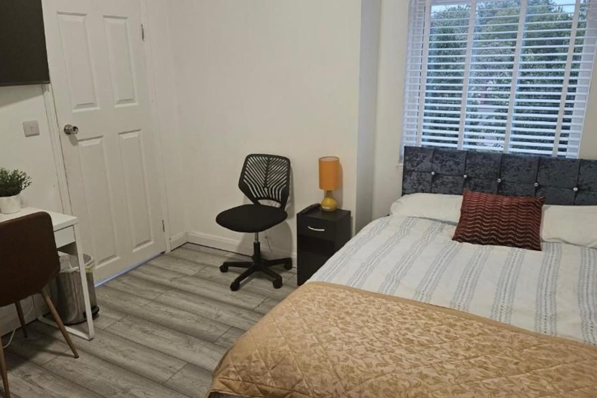 A bedroom with a bed, a desk, a chair, a bedside table with a lamp, and a window with blinds.