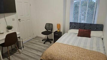 A bedroom with a bed, a desk, a chair, a bedside table with a lamp, and a window with blinds.