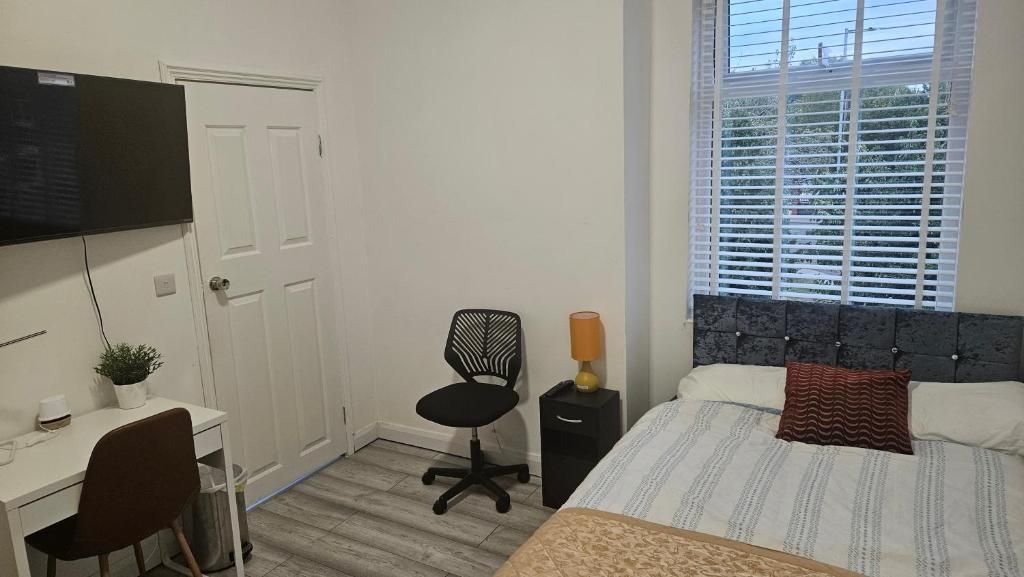 A bedroom with a bed, a desk, a chair, a TV, and a window with blinds.