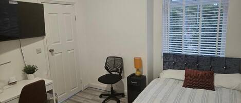 A bedroom with a bed, a desk, a chair, a TV, and a window with blinds.