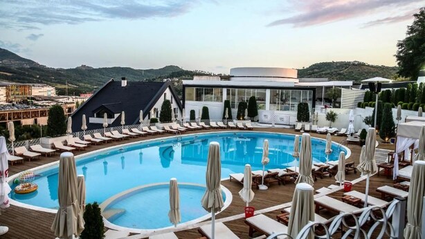 Un espace piscine doté de chaises longues et de parasols, entouré de bâtiments et d’un paysage vallonné.