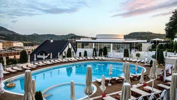 A pool area with lounge chairs and umbrellas, surrounded by buildings and a hilly landscape.
