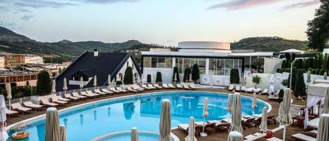 A pool area with lounge chairs and umbrellas, surrounded by buildings and a hilly landscape.