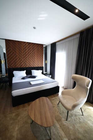 Ein modernes Hotelzimmer mit einem großen Bett, einem runden Holfnachtisch, einem modernen Sessel und einem großen Fenster mit Vorhängen.