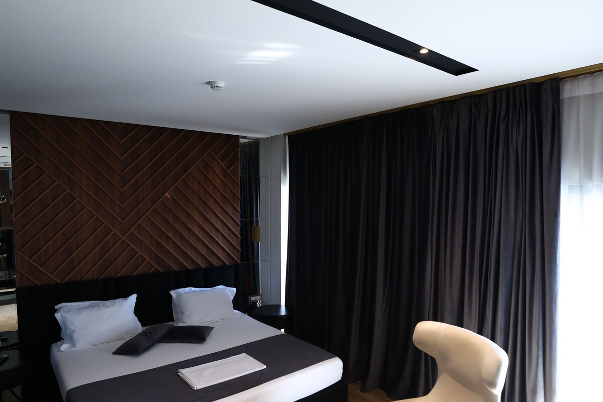 A modern hotel room with a large bed, a bedside table, a chair, and a view of the outdoors.