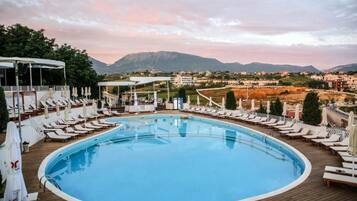 Zona de piscina con sillones de descanso y sombrillas, rodeada por una terraza de madera, con vistas a un paisaje montañoso.