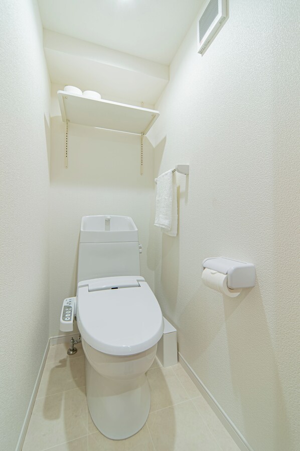 A small, white bathroom with a toilet, a shelf above the toilet, a towel hanging on the wall, and a toilet paper holder.