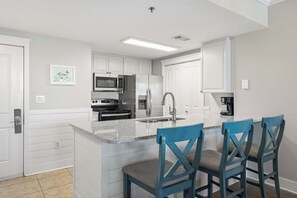 Een moderne keuken met witte kasten, een centraal eiland met een spoelbak, een fornuis en een magnetron. Er staan drie blauwe stoelen bij het eiland.