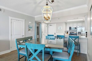 Een eetgedeelte met een glazen tafel en blauwe stoelen, een keuken met witte kasten, en een plafondlamp.