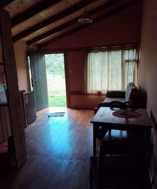 A room with a wooden ceiling, a table with chairs, a window with curtains, and a door leading outside.
