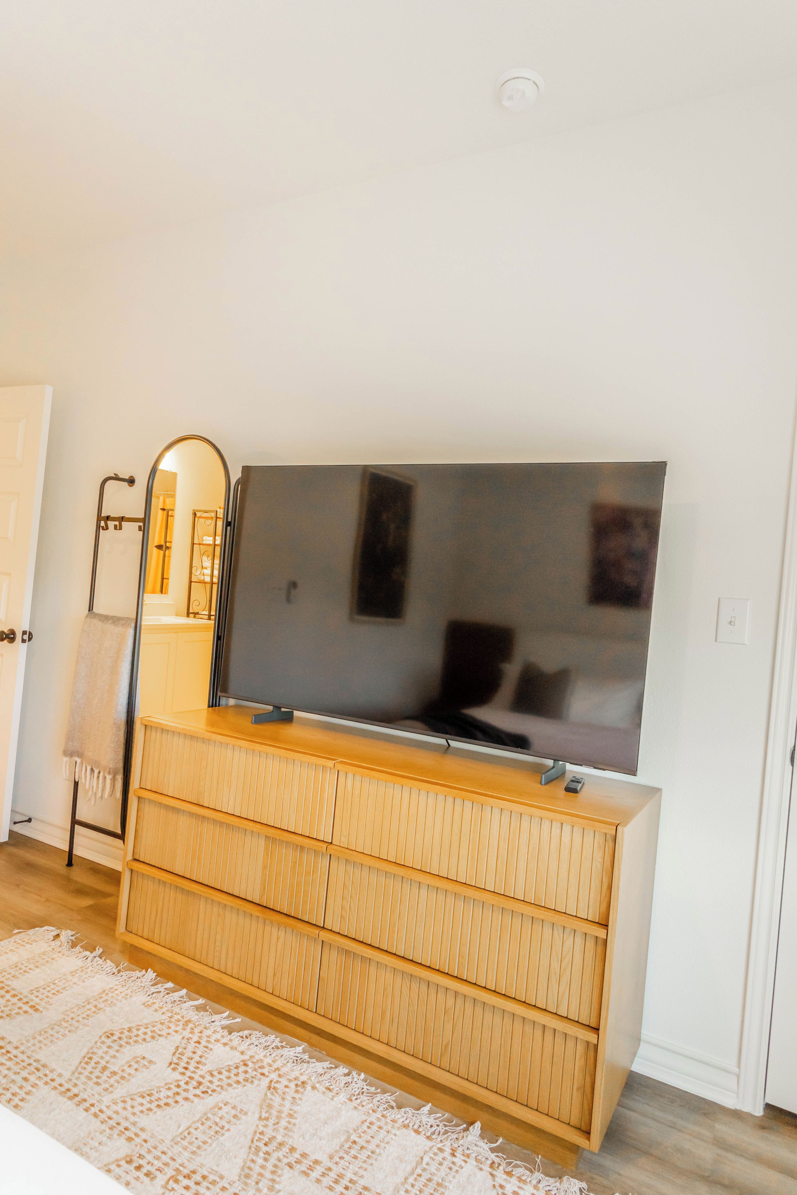A modern living room with a flat-screen TV on a wooden cabinet, a mirror on the wall, and a patterned rug on the floor.