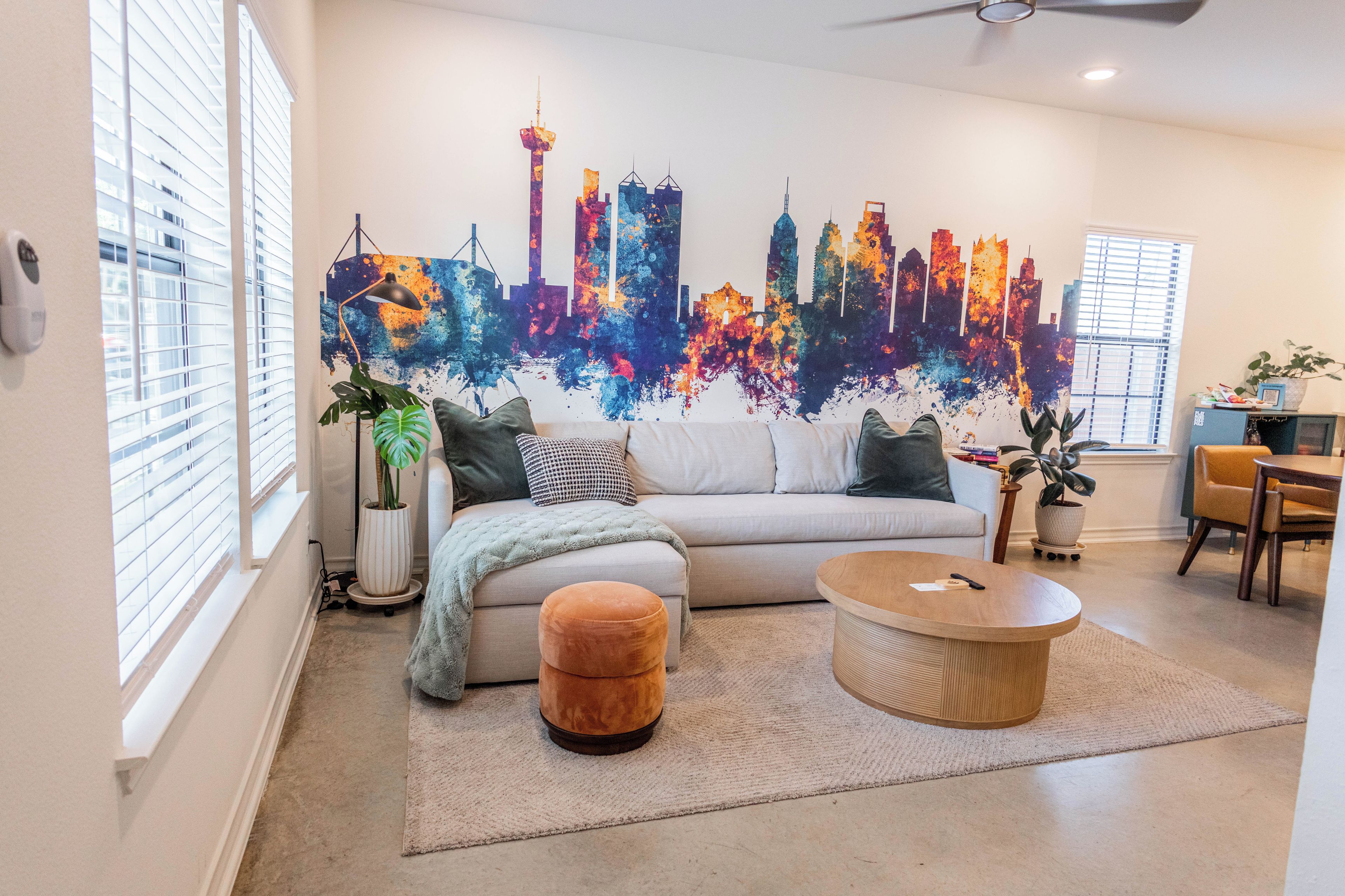 A living room with a sofa, ottoman, coffee table, and a city skyline mural on the wall.