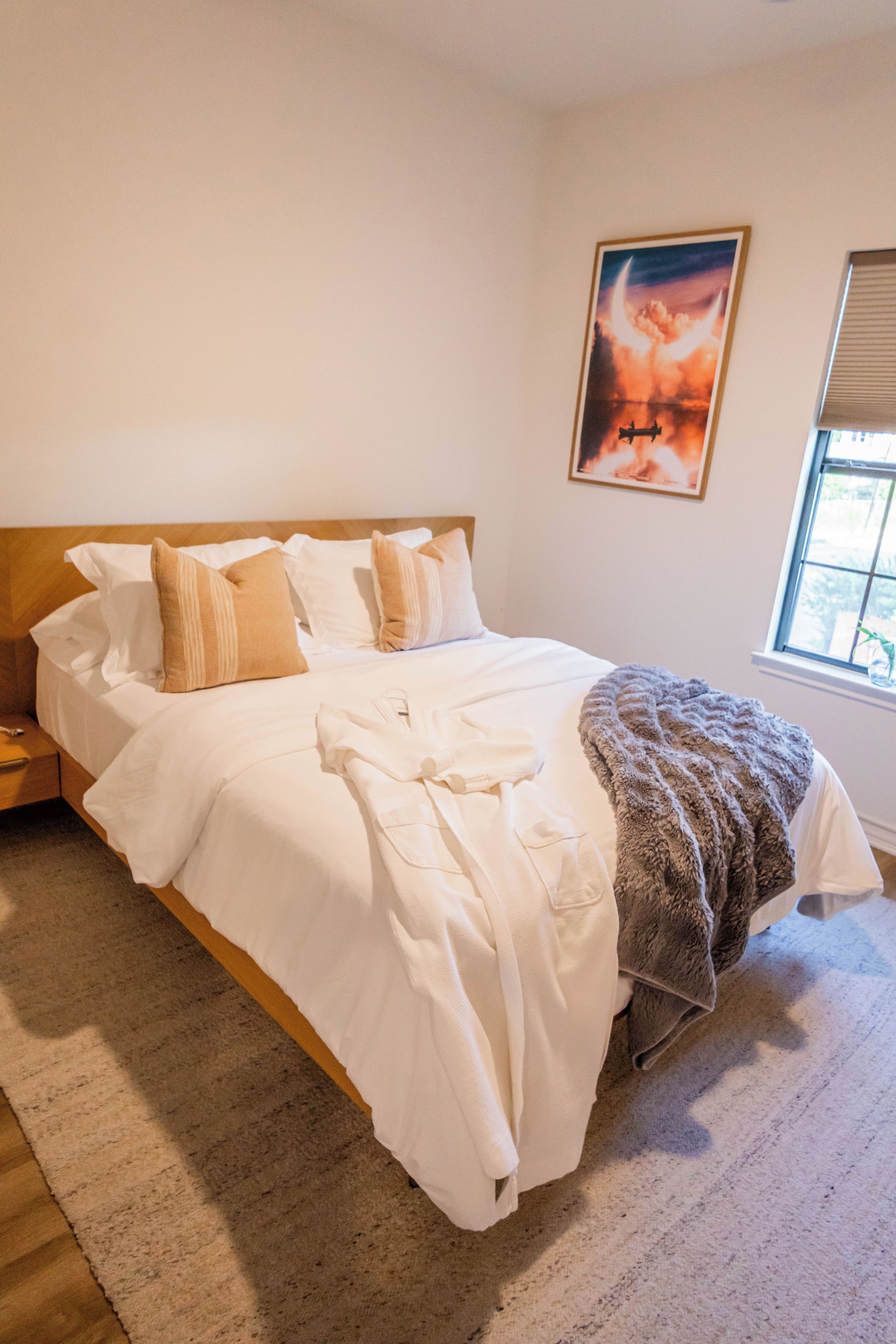 A bedroom with a large bed, a nightstand, a window with blinds, and a framed picture on the wall.