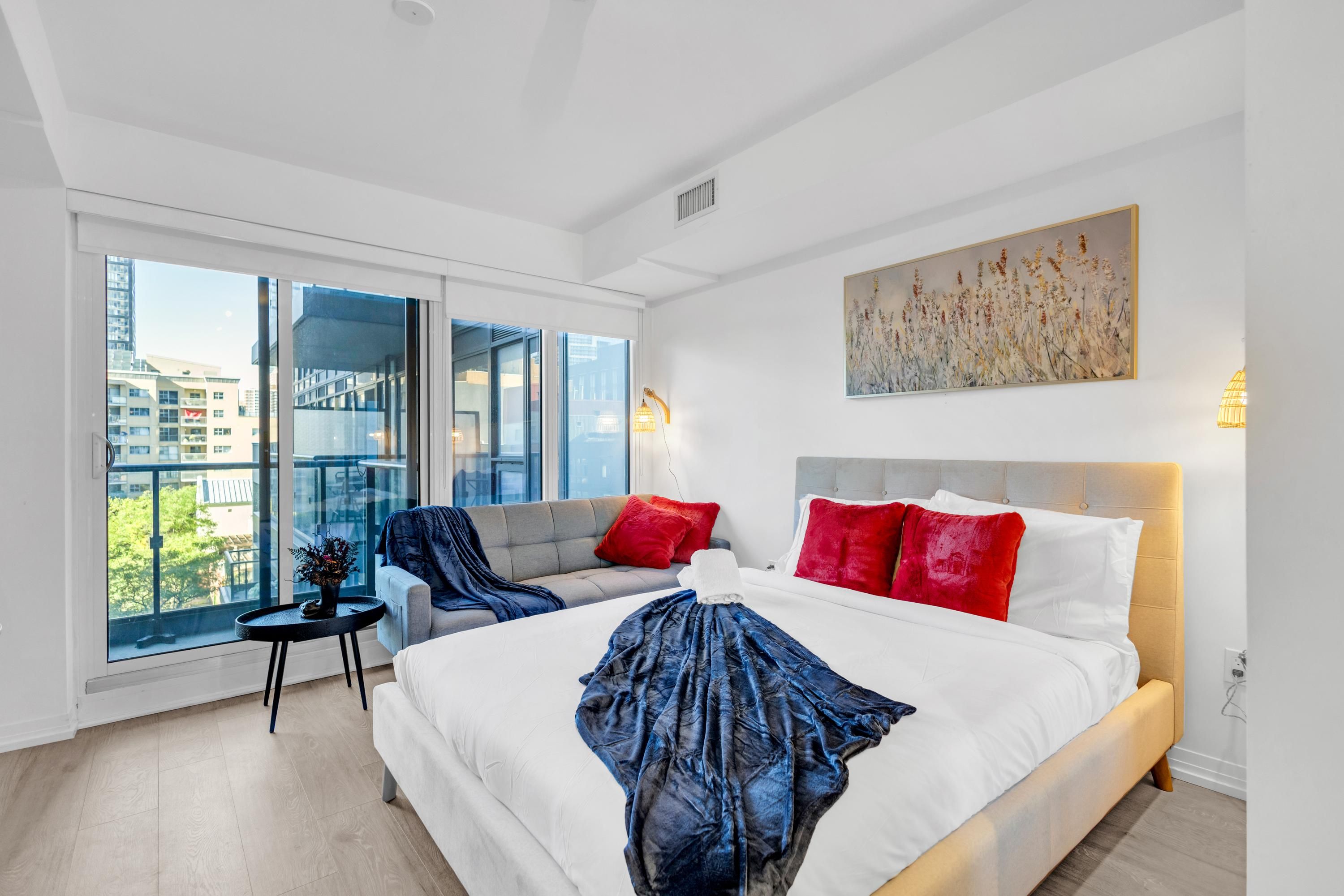 A modern hotel room with a large bed, a seating area with a sofa and armchair, a small table with a vase, and a large window offering a city view.