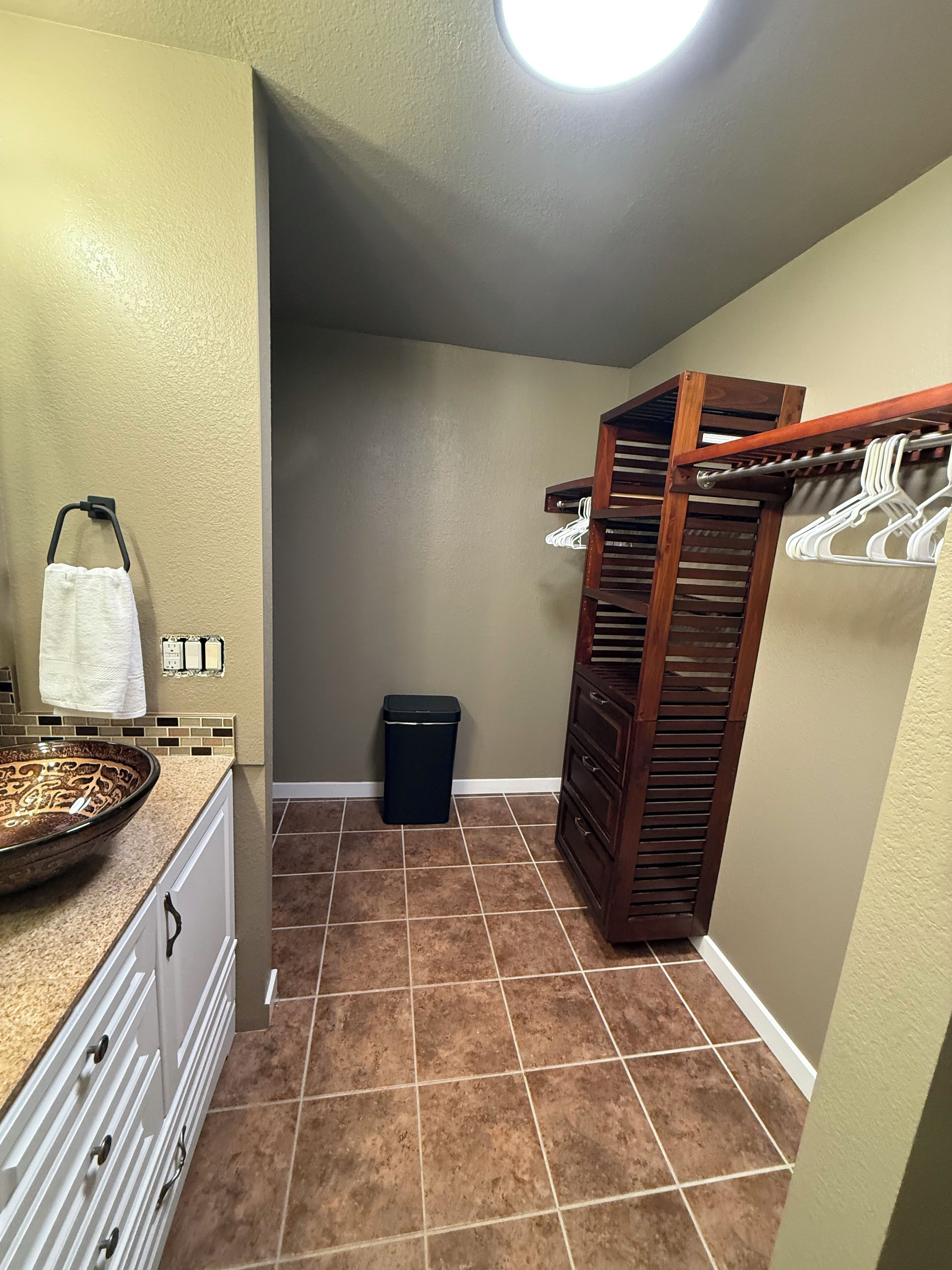 A bathroom with a beige wall, a white towel hanging, a black trash bin, a wooden rack with white hangers, and a brown tiled floor.