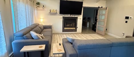 Un salon moderne avec un grand canapé d’angle gris, un téléviseur à écran plat fixé au-dessus d’une cheminée et une table basse en bois.