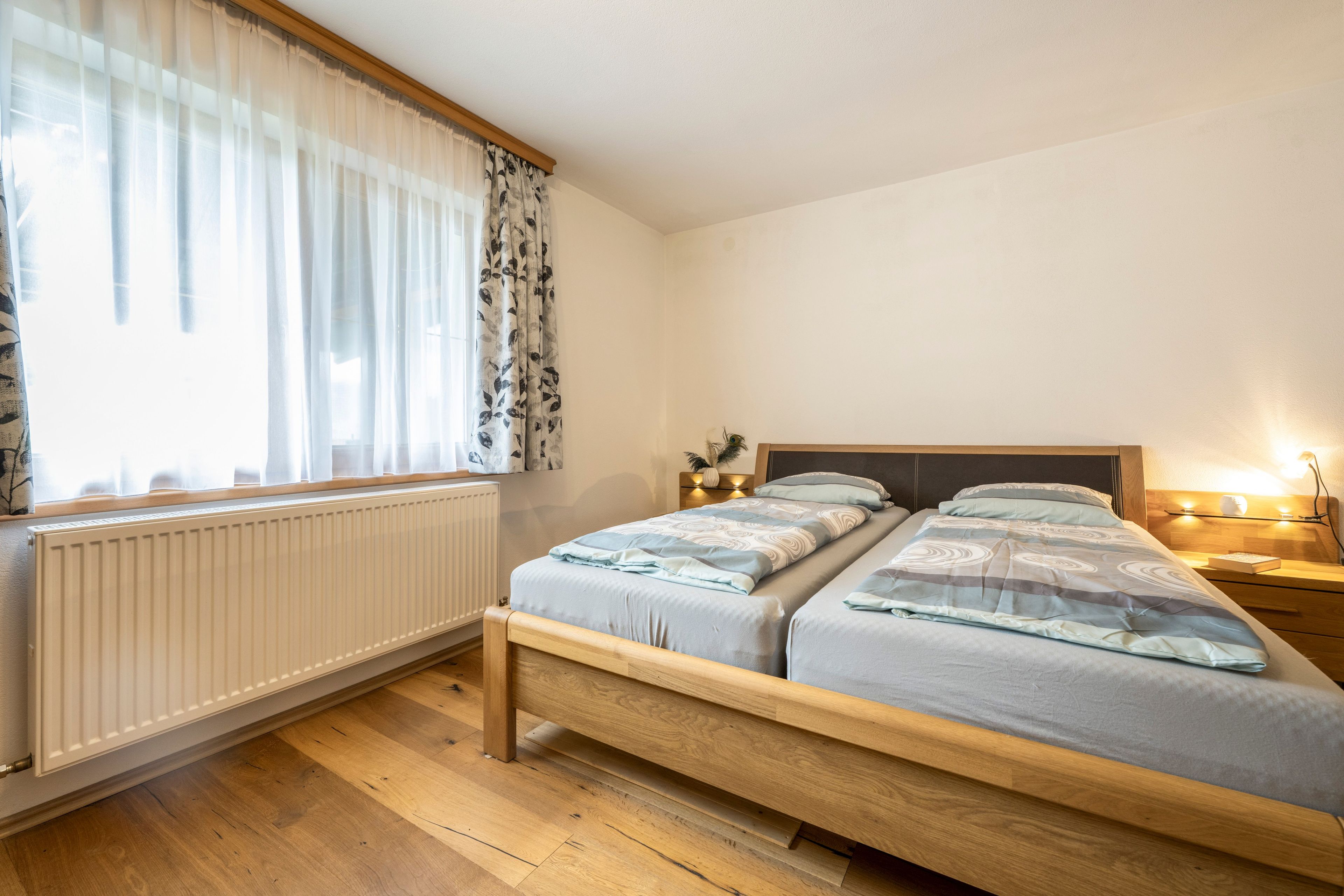 Ein Schlafzimmer mit einem Doppelbett, einem Nachttisch mit Lampe, einem Fenster mit leichtem Vorhang und einer Heizung.