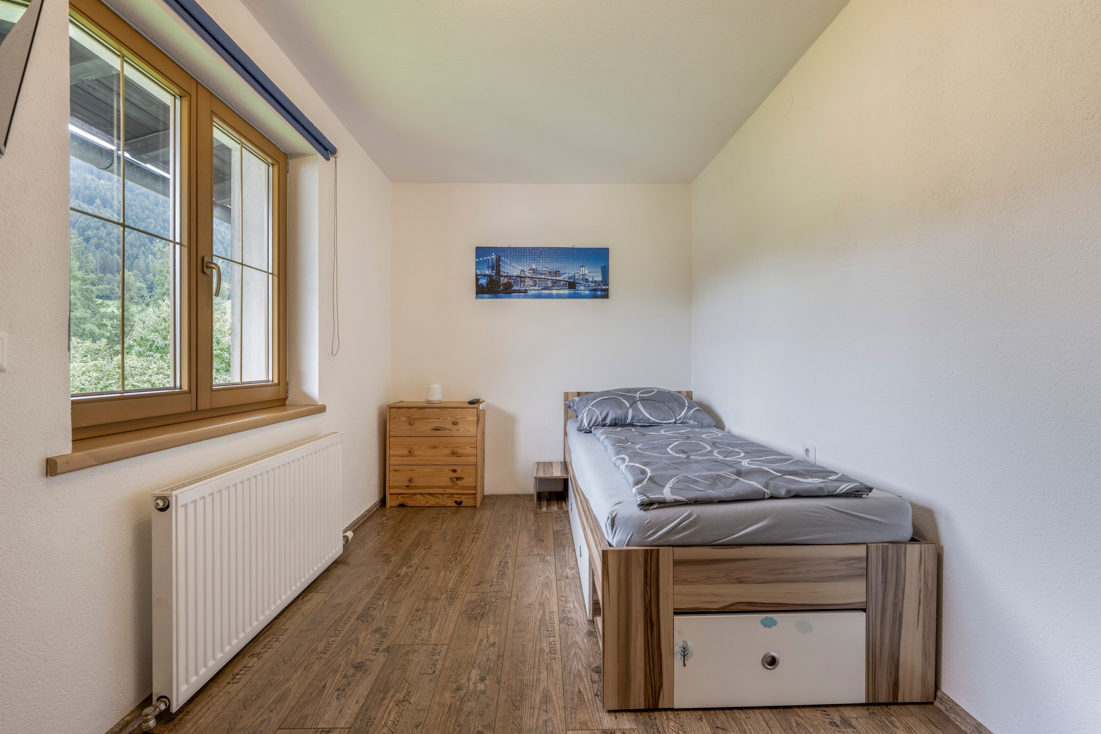 Une petite chambre avec un lit simple, une commode en bois, un radiateur et une fenêtre donnant sur des arbres.