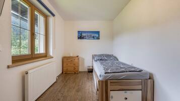 Une petite chambre avec un lit simple, une commode en bois, un radiateur et une fenêtre donnant sur des arbres.