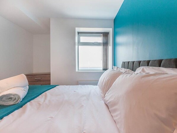 A bedroom with a bed, a window, a nightstand, and a wall with a blue accent. - Private Beach-Front Apartment on the English Riviera with free, private parking (Paignton)