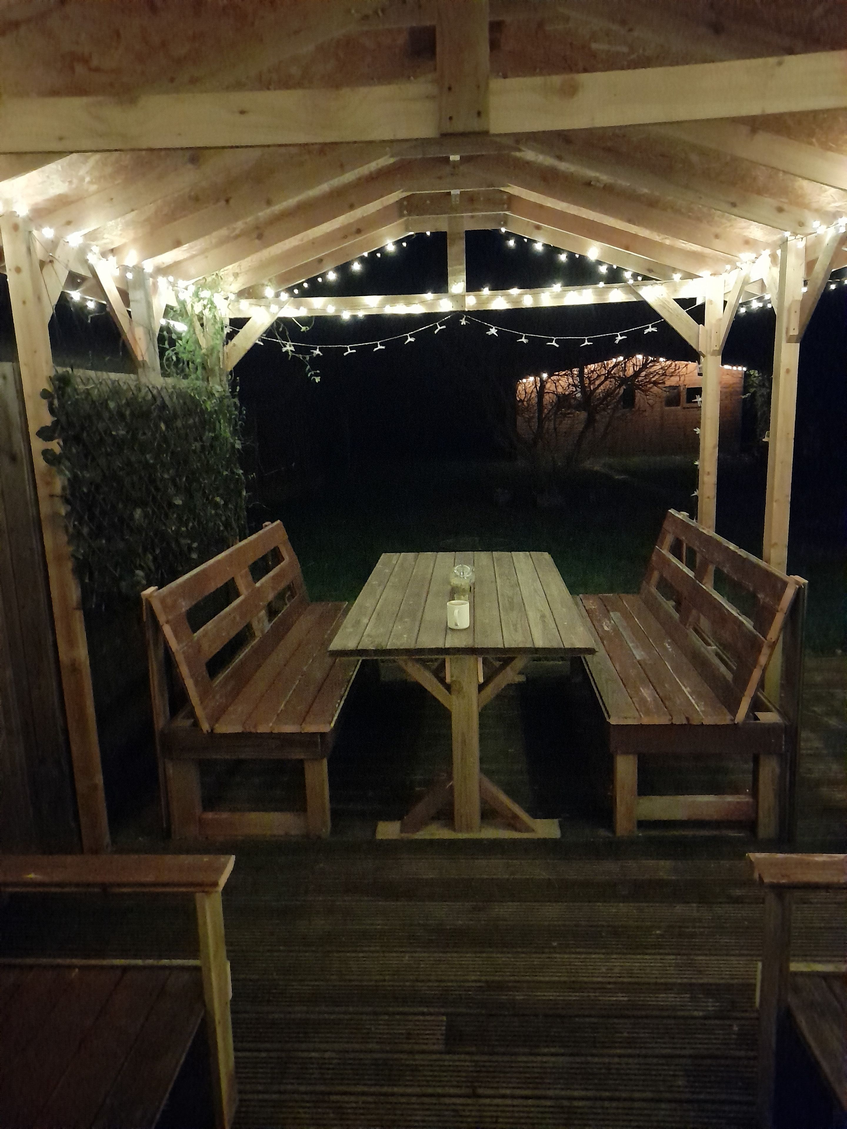 A wooden outdoor patio with a table and benches, string lights overhead, and a hedge to the left.