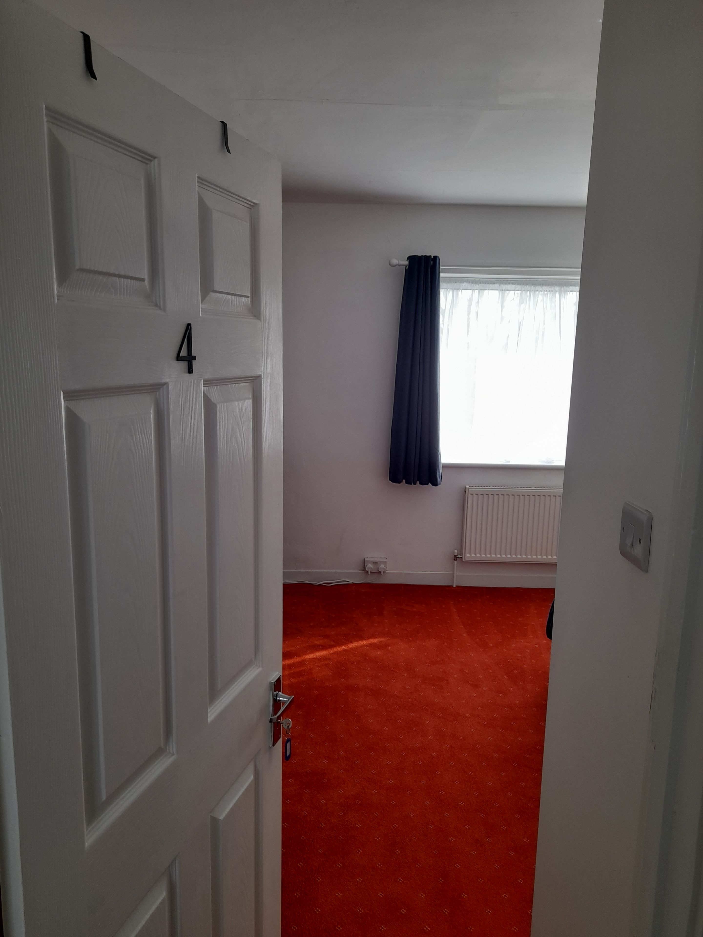 A room with a white door marked with the number 4, a window with a curtain, and a red carpeted floor.