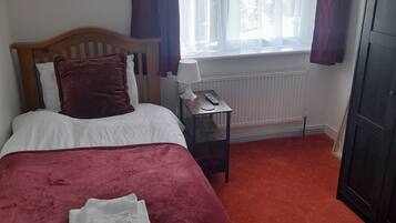 Un dormitorio con cama, una mesita de noche con lámpara, un radiador y una ventana con cortinas.