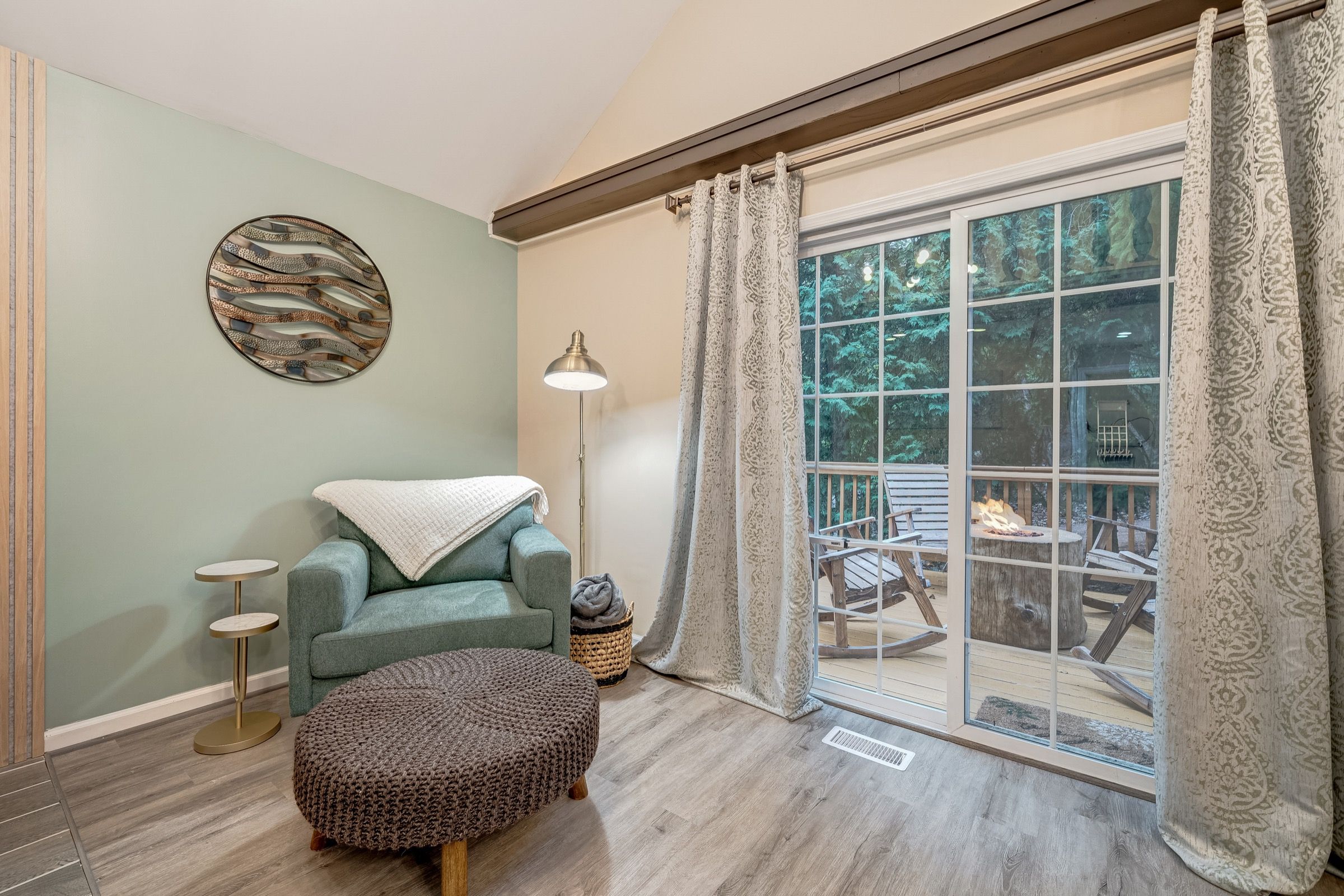 A cozy room with a teal sofa, a round ottoman, a floor lamp, and a large window with curtains.