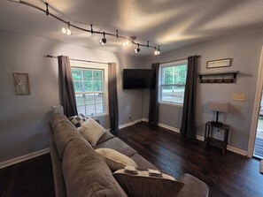 Ein Wohnzimmer mit einer Couch, zwei Fenstern, einem Flachbildfernseher und einer Lampe.